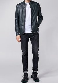 Giubbotto di pelle verde scuro con spalle rigate, indossato sopra una camicia bianca. Abbinato a jeans scuri e scarpe nere con suola verde.