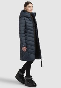 Tengerészkék puffertakó kapucnival és átlós steppeléssel. Teljes hosszúságú cipzárral és oldalsó zsebekkel rendelkezik, fekete hócsizmákkal párosítva.