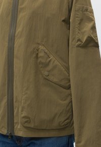 Veste vert olive avec grande poche avant boutonnée affichant le logo "Barbour" et fermeture éclair frontale, portée sur un jean bleu.