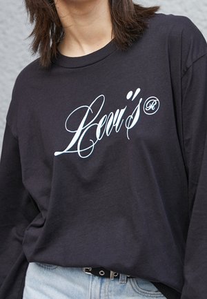 Personne portant un t-shirt noir à manches longues avec "Levi's" en écriture blanche sur la poitrine et un jean bleu clair avec une ceinture noire.