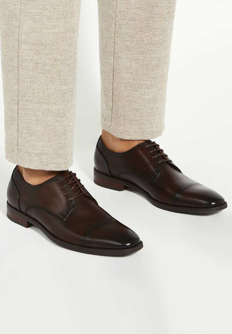 Chaussures habillées en cuir marron avec un embout pointu, un laçage et une finition polie. Exposées contre un pantalon de couleur claire.