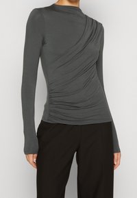 Grå långärmad topp med samlad detalj på ena sidan och hög krage. Tillverkad av mjukt, stretchigt material. Matchas med svarta byxor.