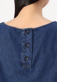 Denim shirt in marineblauw, met een brede halslijn en drie knopen aan de achterkant voor sluiting. De stof lijkt glad en textuur te hebben.