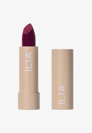 Pomadka ILIA w beżowej tubie z białym brandingiem; ma bogaty burgundowy wkład. Gładka tekstura i klasyczny kształt szminki.