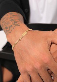 Gold chain bracelet featuring a flat tag with engraved text, showcasing a twisted link design. Skin tone visible beneath the accessory.