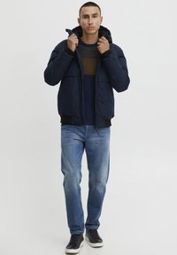 Marineblauwe gewatteerde jas met rits en capuchon, met ribbelmanchetten, gecombineerd met blauwe jeans en zwarte sneakers, tegen een lichte achtergrond.