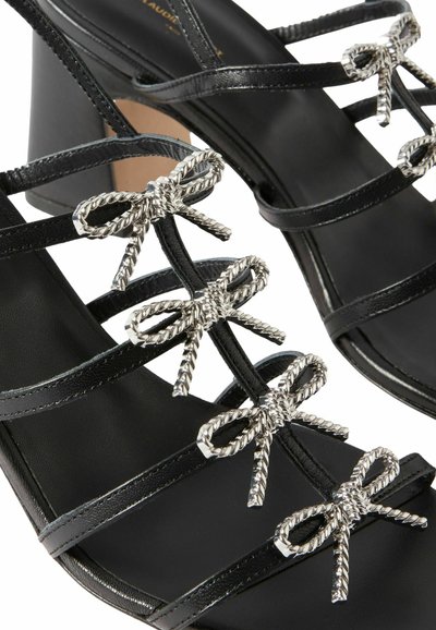 Sandalias negras de tacón con diseño de múltiples tiras que cuentan con detalles de lazos entrelazados plateados. Textura suave y diseño de tacón cónico que complementan el estilo.