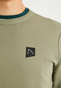 Sudadera verde oliva con un cuello acanalado, que presenta un parche cuadrado negro con un contorno gráfico blanco. Textura suave, diseño casual.