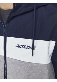 Marinblå och grå zip-up jacka med ett vitt rand och logotyp, dragskohet, mjuk textur och färgblockerad design.