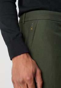 Manica di un pullover scuro accanto a pantaloni verde oliva con cuciture decorative in tre colori: verde scuro, giallo e nero.