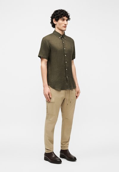 Άνδρας με φουντωτά μαλλιά, που φοράει πουκάμισο με κοντά μανίκια σε ελιά χρώμα, μπεζ παντελόνι cargo και σκούρα καφέ μποτάκια που φτάνουν στον αστράγαλο, στέκεται μπροστά σε απλό φόντο.
