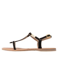 Black suede flat sandal with a T-strap design, adjustable ankle buckle, and a tan rubber sole, featuring stitched detailing.