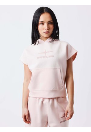 Femme aux cheveux noirs et raides portant un sweat à capuche sans manches rose pâle et un pantalon de survêtement assorti avec un logo devant, debout devant un fond clair.