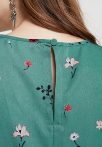 Tessuto verde con motivo floreale, dotato di chiusura a bottone posteriore e dettaglio a chiave. Presenta piccoli fiori rossi, rosa e blu su una texture liscia.