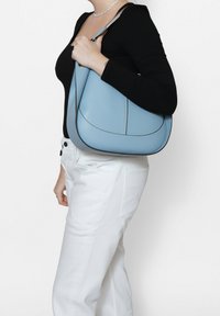 Borsa a spalla in pelle azzurro chiaro con forma curva, cuciture scure a contrasto e una sola tracolla, tenuta contro un top nero e pantaloni bianchi.
