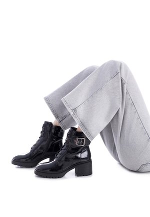 Botas negras de charol hasta el tobillo con un tacón grueso, cordones en la parte delantera, cremallera lateral y detalle de hebilla. Llevadas con pantalones de pierna ancha en gris claro.