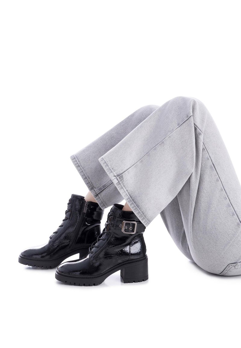 Botas negras de charol hasta el tobillo con un tacón grueso, cordones en la parte delantera, cremallera lateral y detalle de hebilla. Llevadas con pantalones de pierna ancha en gris claro.