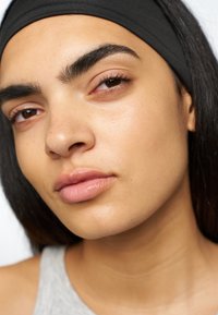 Nærbilde av et ansikt med glatt, lys brun hud, veldefinerte bryn og dempede rosa lepper. En svart hårband rammer inn hårfeste.