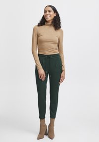 Hřejivý krémový rolák s dlouhým rukávem je kombinován se zelenými jogger kalhotami s šňůrkovým pasem a dvěma předními kapsami. Modelka má na sobě béžové boty na podpatku.