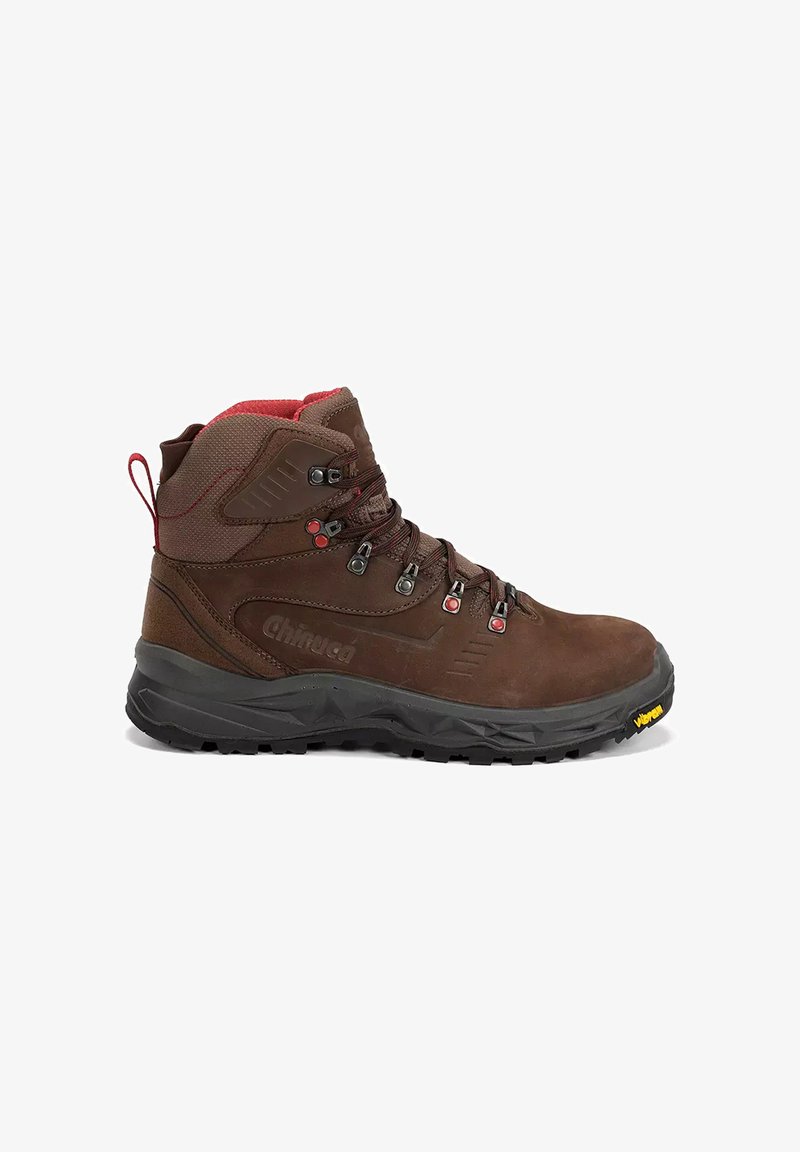 Botte de randonnée marron à coupe mi-haute avec des accents rouges, des œillets métalliques, un col rembourré, une semelle noire robuste et un logo Vibram sur le bord avant.