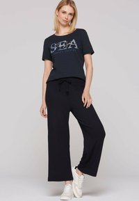 Camiseta gráfica navy con estampado "SBA Sunset Sailing", combinada con pantalones negros de canalé sueltos; modelo usando zapatillas blancas.