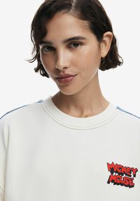 Felpa bianca con dettagli azzurri, caratterizzata da scritte rosse ricamate "Mickey Mouse". Tessuto liscio, collo rotondo e cuciture visibili.