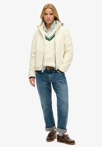 Witte pufferjack met een zwarte rits, gelaagd over een crèmekleurige kabelgebreide trui en een groene gestreepte colbert; blauwe jeans met opgerolde manchetten en bruine schoenen.