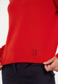 Pull en maille rouge avec un ourlet côtelé, arborant un "IF" noir brodé en bas à gauche. Texture lisse, manches longues.