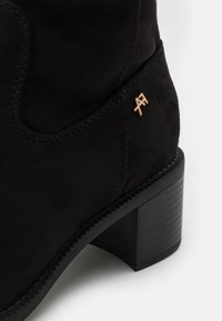 Black suede ankle boot with a block heel, featuring a gold logo accent and detailed stitching along the seams. Smooth texture.