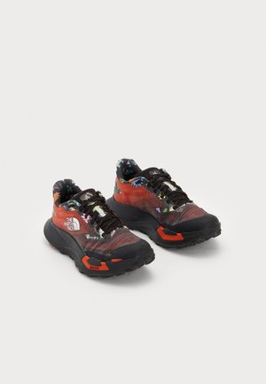 Chaussures de trail avec un dessus en mesh rouge et noir, des accents floraux colorés, une épaisse semelle noire et des détails réfléchissants sur les lacets.