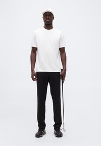 Άνδρας με λευκό καπέλο, λευκό κοντομάνικο πουκάμισο, μαύρο παντελόνι και παπούτσια γκολφ κρατώντας ένα γκολφ μπαστούνι μπροστά σε απλό λευκό φόντο.