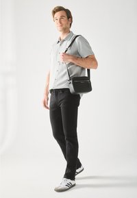 Sac à bandoulière noir avec une surface texturée, poche zippée à l'avant et bandoulière réglable, associé à une chemise gris clair et un jean noir.