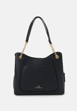 Schwarze Handtasche aus Kunstleder mit geschwungener Form, goldfarbener Kettenakzenten, einer Fronttasche und geprägtem Logo-Detail am Boden.