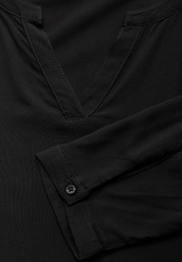 Blusa nera con scollatura a V, maniche lunghe con polsini abbottonati e tessuto liscio e strutturato con dettagli di cucitura in contrasto.