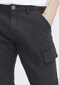 Pantaloni cargo neri con tasca laterale a soffietto e chiusura con bottone frontale, indossati da una persona con una mano in tasca su uno sfondo neutro.