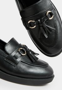 Schwarze Lederloafers mit goldenen Beschlägen, die Quasten und eine erhöhte Sohle aufweisen. Glatte Textur, klassisches Design und runde Zehenform.