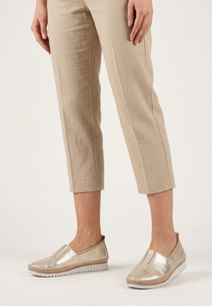 Person wearing beige cropped pants and metallic slip-on casual shoes, standing on a plain light background.