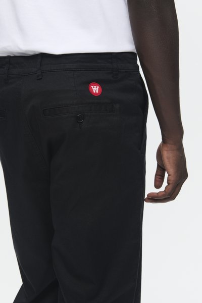 Pantalones de algodón negros con un parche de logo rojo en el bolsillo trasero, cierre de botón y silueta ajustada. Textura suave visible.