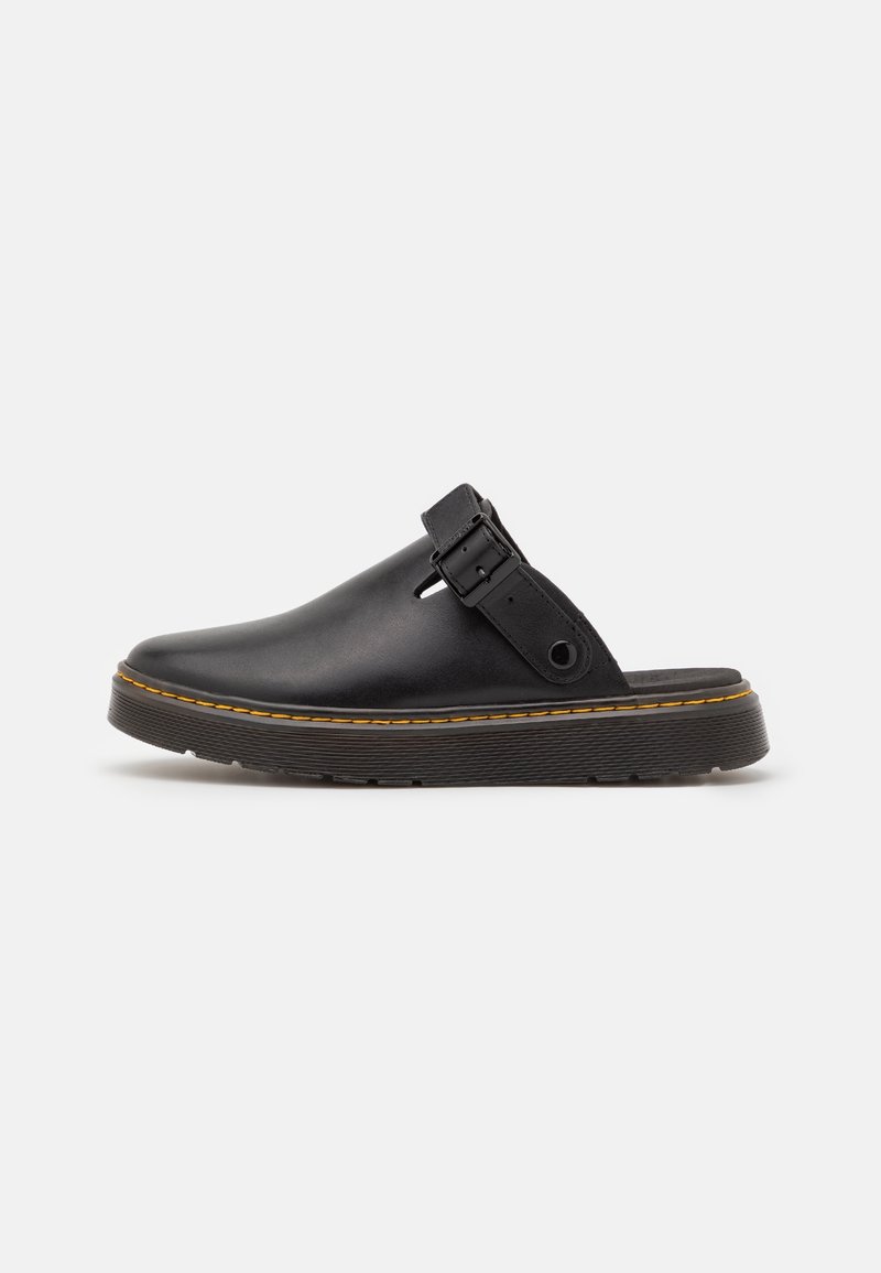 Chaussure en cuir noir à enfiler avec semelle épaisse, surpiqûres jaunes et bride ajustable avec boucle sur un fond clair uni.