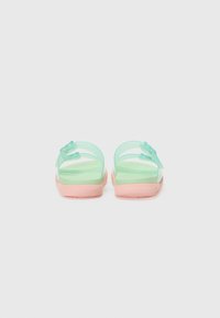 Grønne transparente sandaler med en rosa såle, og to blomsterformede detaljer på remmene. Teksturert fotseng for godt grep. Bakfra.
