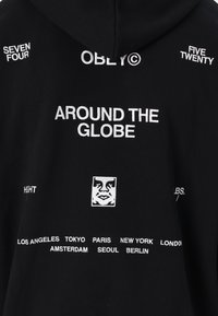 Schwarze Kapuzenjacke mit weißem gesticktem Text, der "AROUND THE GLOBE", Städtenamen und grafischen Details eines Gesichts enthält.