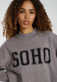 Grå tröja med hög halsringning, med "SOHO" i fet svarta bokstäver. Mjuk textur, lös passform och sänkta axelsömmar.