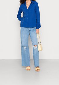 Blaues Hemd mit langen Ärmeln, kombiniert mit hellblauen, weit geschnittenen, ripped Jeans. Die Accessoires umfassen eine kleine gelbe Handtasche und gelbe Sandalen.