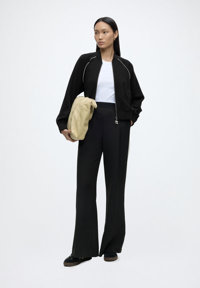 Femme debout, portant une veste noire zippée, une chemise blanche, un pantalon noir à jambes larges, des chaussures noires, tenant un sac beige sous le bras.