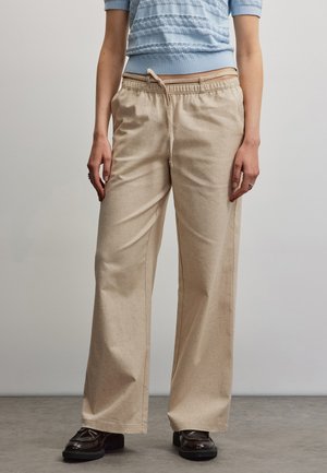Kvinde iført beige vidde bukser med snøre i taljen, lyseblå tekstureret kortærmet top og sorte loafers, stående på en grå gulv.