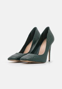 Une paire de chaussures à talons aiguilles vert foncé avec des bouts pointus et un motif en cuir de serpent texturé sur fond blanc.