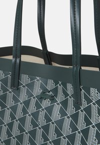 Borsa a tracolla verde realizzata in materiale sintetico testurizzato, con motivi geometrici bianchi e un piccolo logo impresso. Inclusi due manici.