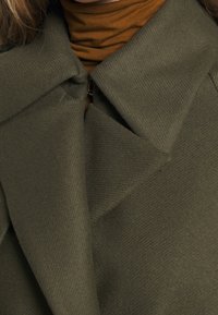 Abrigo verde oliva de tela texturizada y cuello prominente, con cierre de cremallera y escote en capas con un cuello de tortuga marrón debajo.