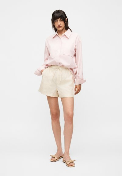 Γυναίκα στέκεται φορώντας ένα ανοιχτό ροζ πουκάμισο με μακριά μανίκια, μπεζ σορτς με κορδόνι και μπεζ σανδάλια με λεπτομέρειες φιόγκων σε απλό φόντο.