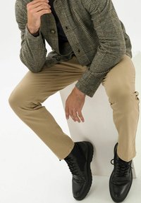 Brązowa kurtka w kratę, czarna koszulka, beżowe spodnie oraz czarne buty sznurowane. Kurtka posiada teksturowany wzór i zapięcie na guziki.